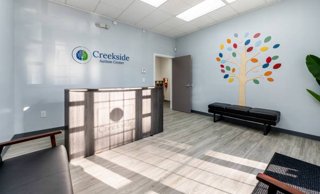 A well lit front desk and waiting area provides a comforting space for parents and children. Creekside Autism logo and tree decoration are present on the walls. Office of Creekside Autism Center